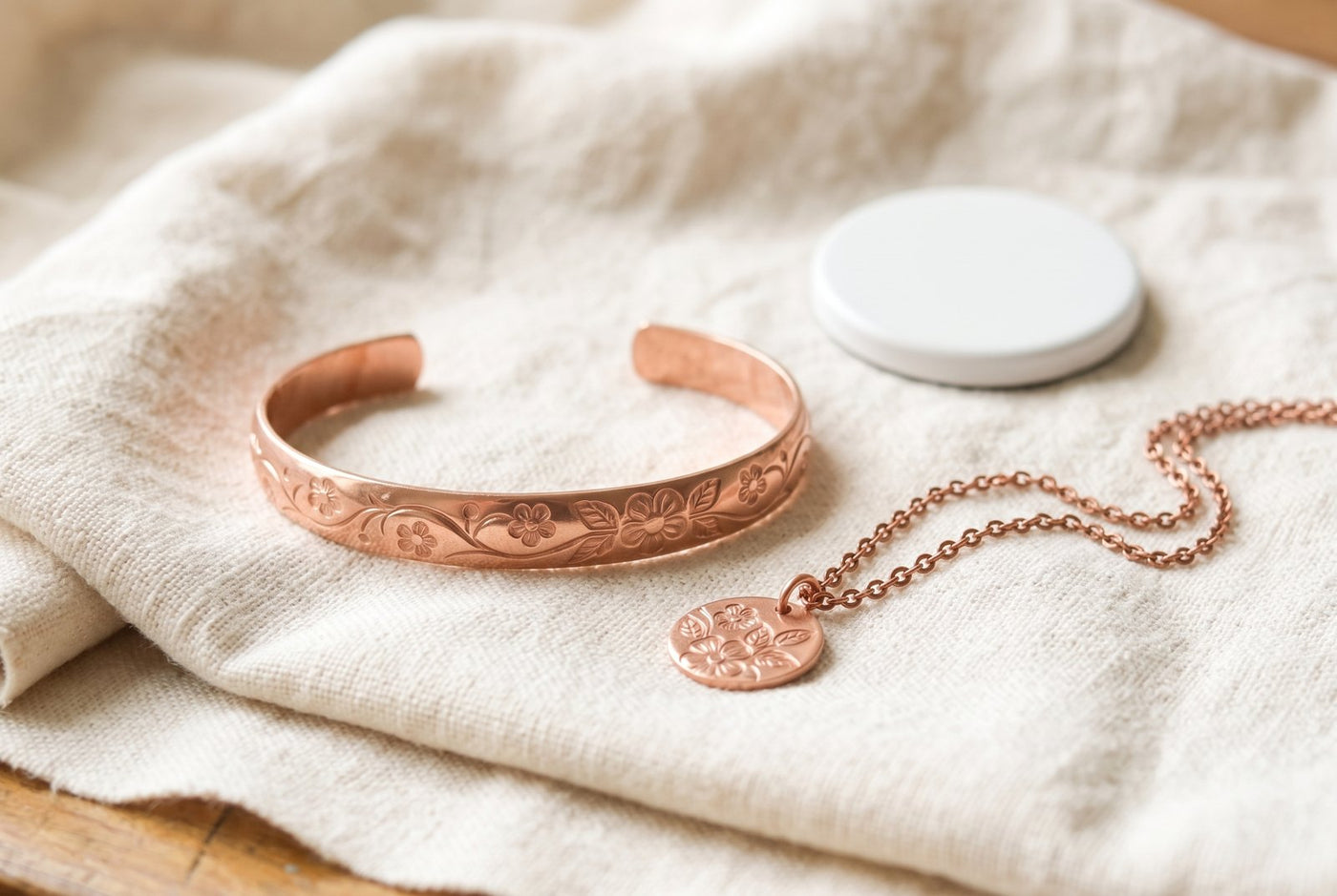 A spread of solid copper pieces on white linen in soft daylight: a cuff engraved with a floral vine, a plain bracelet, a